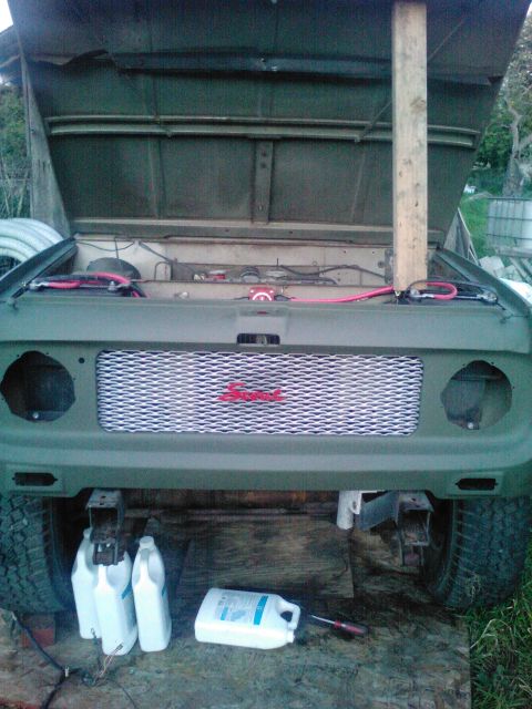 1991 Ford Bronco - The front of a green vehicle with its hood open, revealing engine components and wiring. A custom silver grille with red lettering is visib…