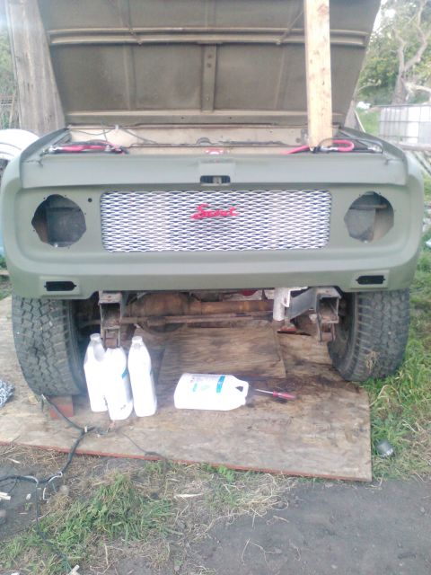 1991 Ford Bronco - The front end of a vintage green SUV is shown with its hood open, revealing a custom silver mesh grille with a red emblem. The vehicle is p…