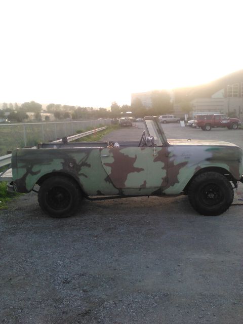1991 Ford Bronco - A side profile of a vintage convertible SUV with a camouflage paint job is parked on a gravel lot. The vehicle has black wheels and a roll…
