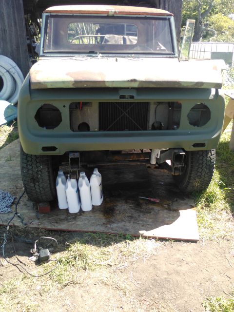1991 Ford Bronco - Front view of a vintage truck with camouflage paint on the hood and fender, undergoing restoration. Four white plastic jugs are lined up in…