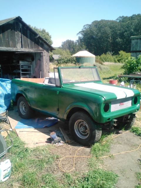 1991 Ford Bronco - A green convertible SUV with white racing stripes on the hood is parked on a dirt and grass area next to a weathered wooden shed.
