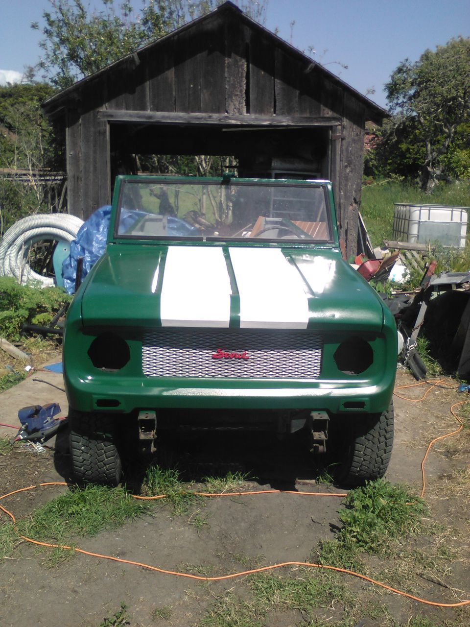 Green 1991 Ford Bronco with white hood stripes and silver grille parked outside a wooden shed.