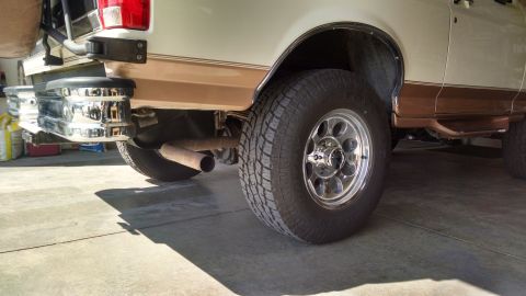 1994 Ford Bronco - The rear passenger side of a tan and white Ford Bronco is shown, focusing on the tire, exhaust pipe, and chrome bumper. The vehicle is park…