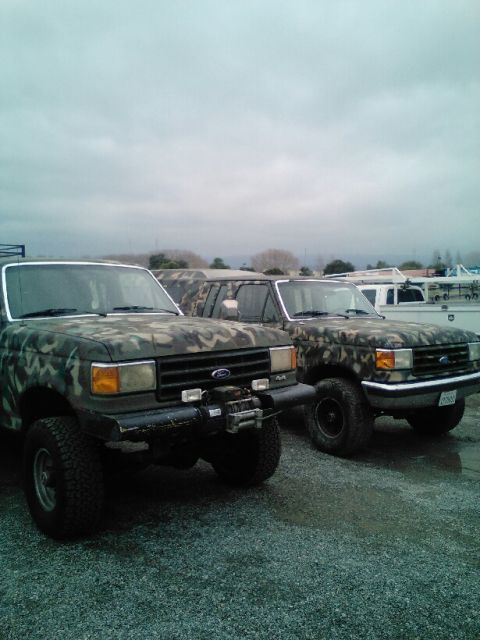 1991 Ford Bronco - Two Ford trucks with camouflage paint are parked on a gravel lot under a cloudy sky. The truck in the foreground has a winch mounted on its…
