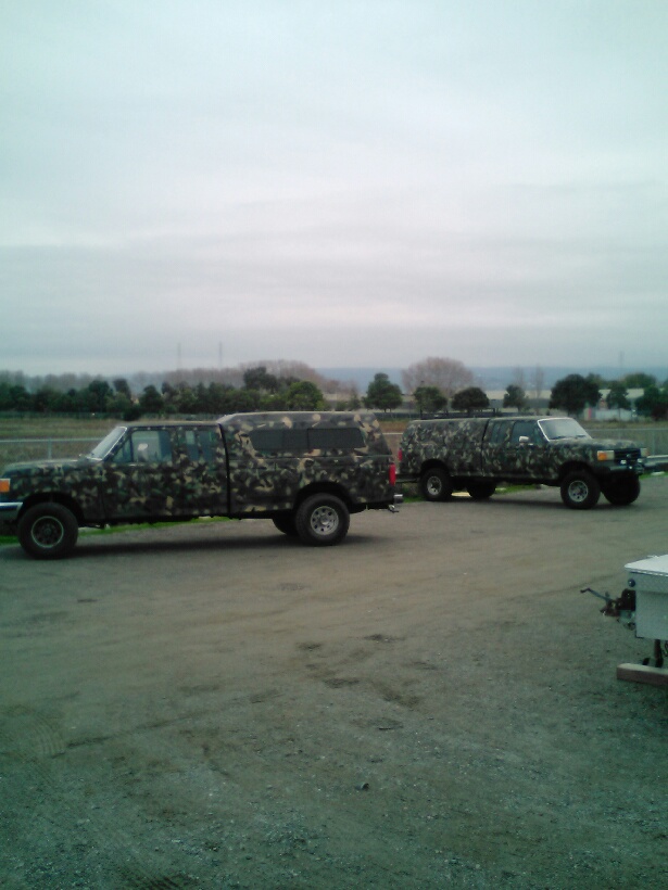 Two camouflage pickup trucks with camper shells parked on a gravel lot.