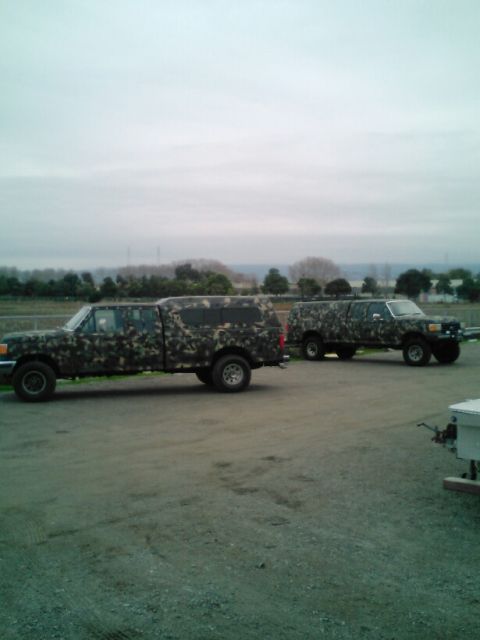 1991 Ford Bronco - Two pickup trucks with camouflage paint are parked on a gravel lot with a wooded area and cloudy sky in the background.
