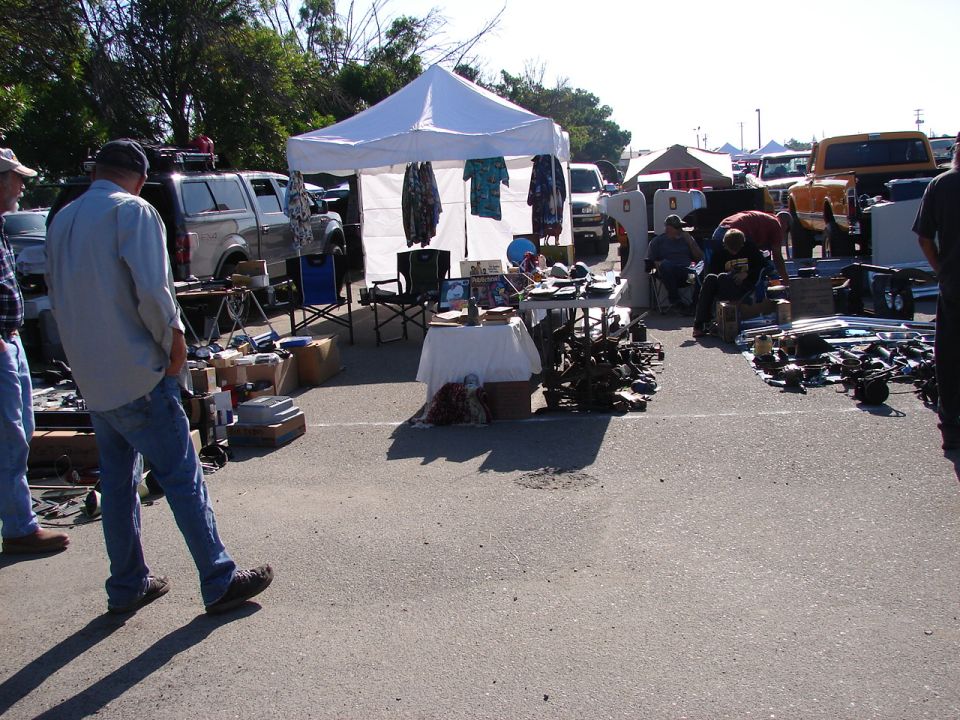 Outdoor market with a white tent displaying clothing and car parts for sale.