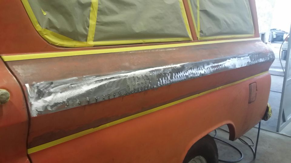Side view of an orange vintage Chevrolet Suburban with body repair on the lower side panel.