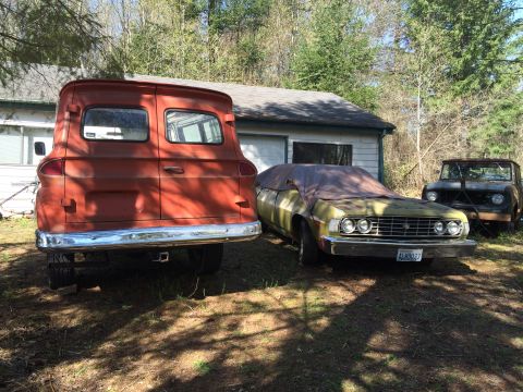 1962 Chevrolet Suburban - The rear of a vintage, orange panel van is parked in a grassy yard next to a garage. A light-colored sedan covered by a tarp and a dark-col…