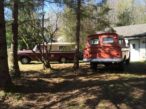 1962 Chevrolet Suburban - A red 1960s panel van is parked next to a maroon pickup truck in a wooded area. The panel van is viewed from the rear, showing its chrome b…