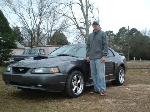 2003 Ford Mustang - A man stands next to a dark gray Ford Mustang, which has chrome wheels and a prominent hood scoop.