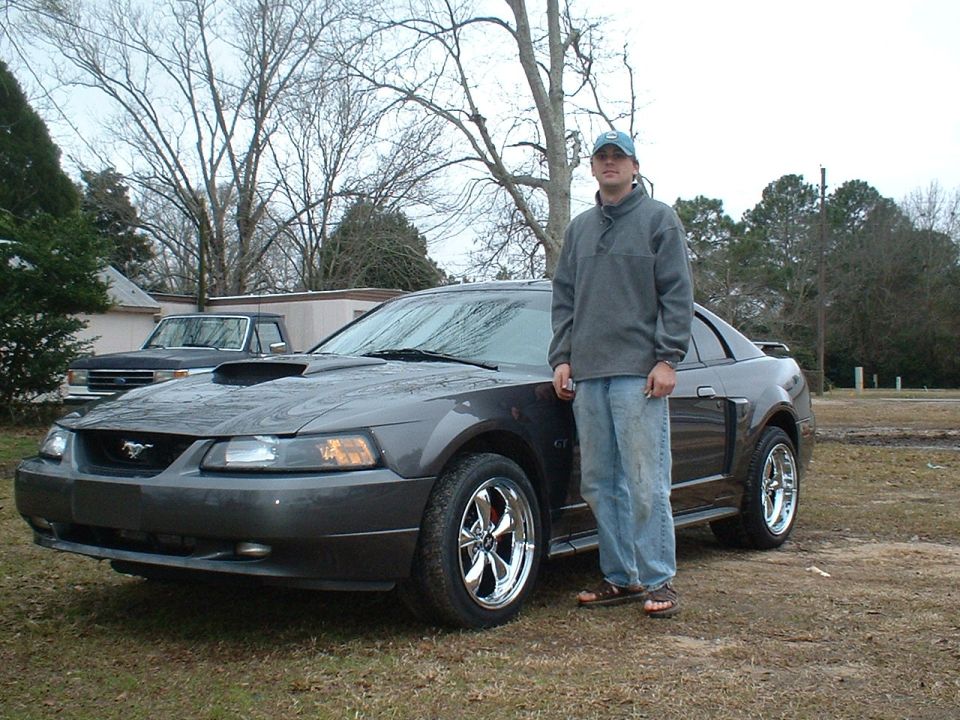 Dark gray Ford Mustang with chrome wheels and a hood scoop, parked on grass with a man standing beside it