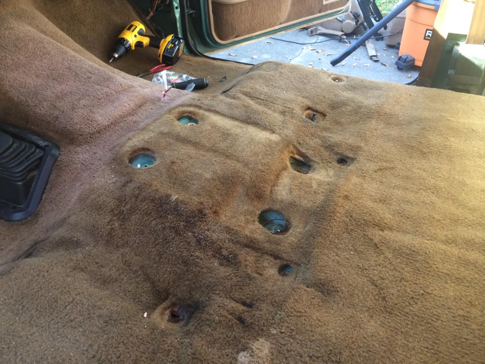 Vehicle interior floor with carpet removed, showing mounting holes and a drill