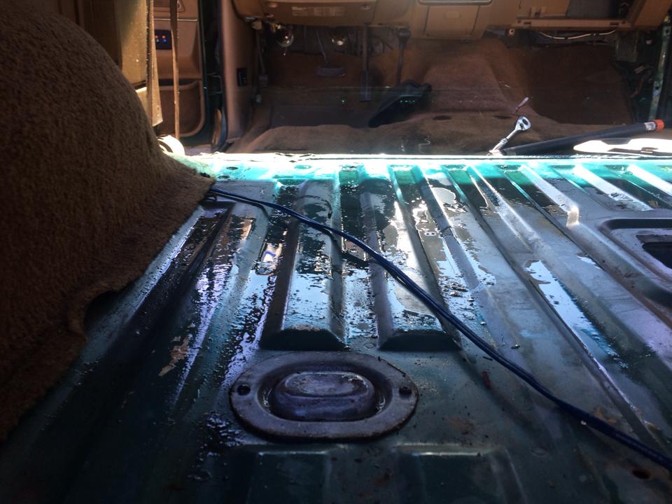 Vehicle interior with carpet removed, showing a wet, green metal floor with tie-down points and wiring