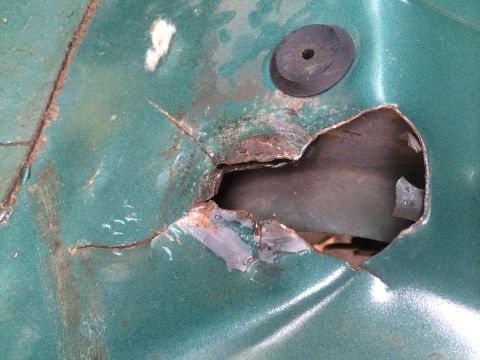 1990 Ford Bronco - Close-up of a rusted hole in a green metal panel, with a black rubber grommet visible.