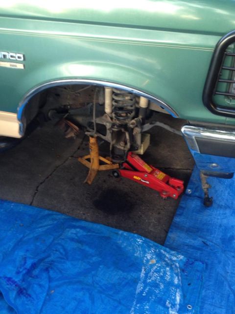 1990 Ford Bronco - The front suspension of a green 1990 Ford Bronco is visible, supported by jack stands and a hydraulic jack.