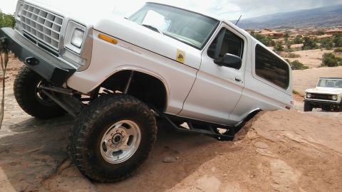 1979 Ford Bronco - Two white SUVs traverse a rocky, sandy landscape with desert vegetation in the background.