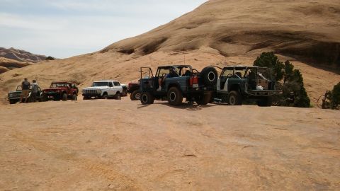 1979 Ford Bronco - Multiple off-road vehicles are parked on a sandy, rocky terrain under a bright sky.