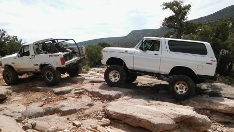 1979 Ford Bronco - Two lifted Ford Broncos navigate a rocky off-road course under a partly cloudy sky.