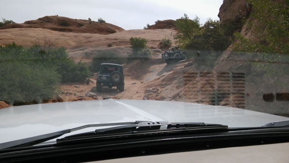 Two off-road vehicles driving on a dirt path in a rocky, desert-like area. View from inside a vehicle