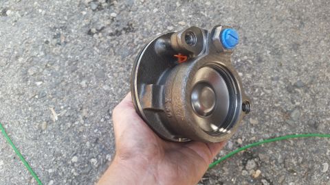1981 Ford Bronco - A brake caliper part is held in a hand over a gravel surface. The part shows internal components and a blue cap on a bolt.