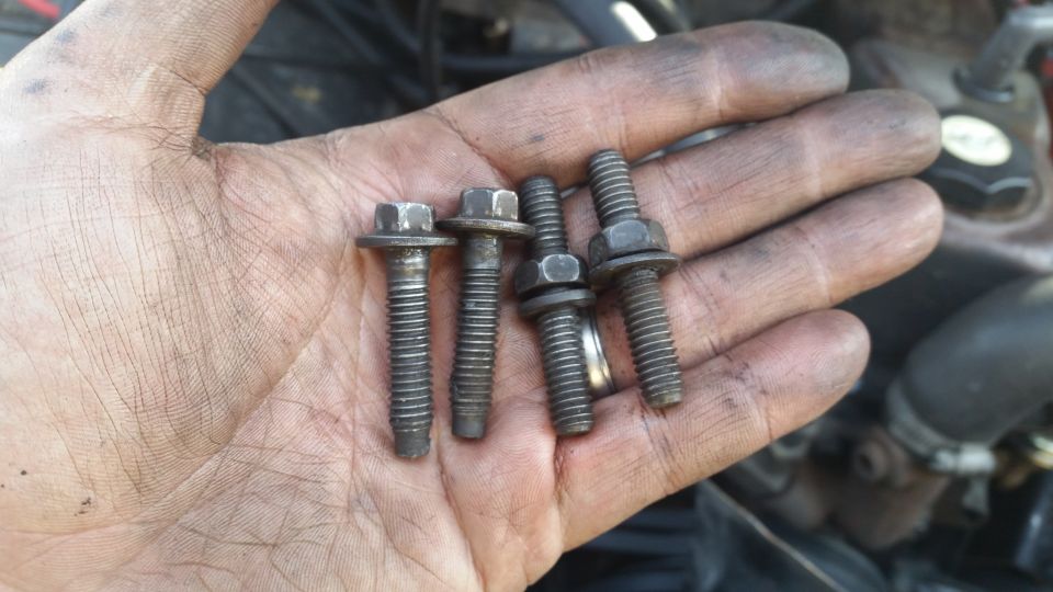 Dirty hand holding four bolts, two with washers, against a blurred engine background