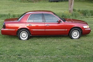 1999 Mercury Grand Marquis - Side view of a red 1999 Mercury Grand Marquis with tinted windows, parked outdoors on a grassy area.