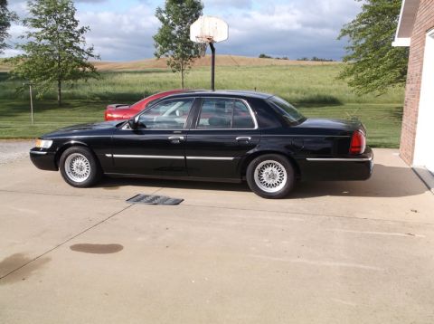 1999 Mercury Grand Marquis - A black sedan is parked on a concrete driveway next to a brick building, with a basketball hoop in the background.