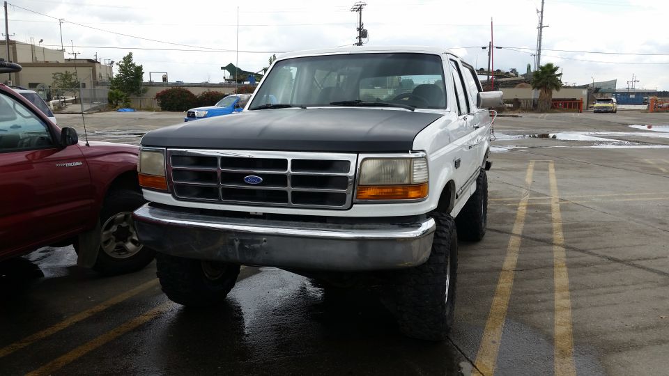 Lifted white Ford Bronco with black hood and oversized tires in a wet parking lot