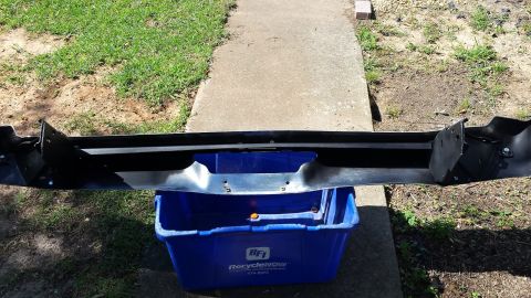 1976 Ford Ranchero - Black painted sheet-metal valance or core support panel laid horizontally across a blue BFI recycle bin on a backyard walkway.