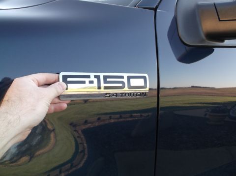 2005 Ford F-150 - A hand holds an F-150 5.4 Triton badge against the fender of a dark blue truck.