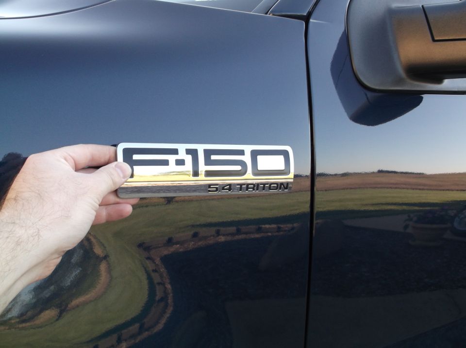 Dark blue Ford F-150 5.4 Triton badge being held by a hand on the vehicle's fender