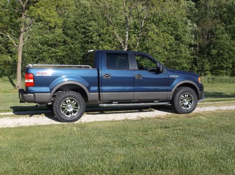 2005 Ford F-150 - A blue pickup truck with custom wheels is parked on a gravel driveway next to a grassy area.