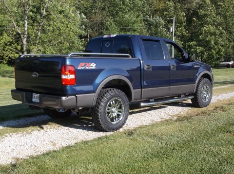 2005 Ford F-150 - This blue Ford F-150 pickup truck is shown from the rear quarter angle, parked on a gravel driveway with trees in the background.
