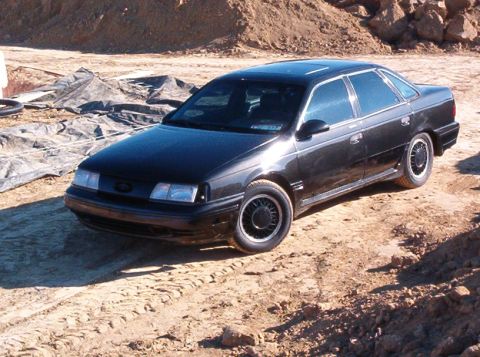 1990 Ford Taurus - A black sedan with a sunroof is parked on a dirt lot, viewed from a three-quarter front angle.