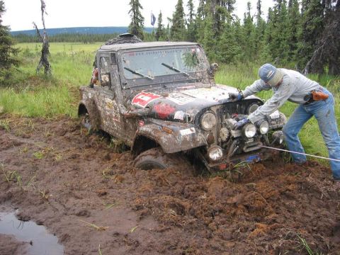 1985 Ford Bronco - Off-road vehicle deeply stuck in mud, with a person attempting to use a winch.