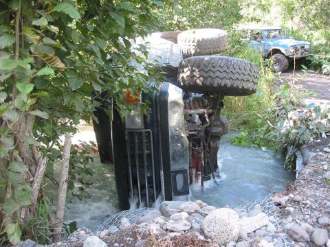 1985 Ford Bronco - An overturned dark SUV is submerged in a stream, while a blue SUV sits on a nearby dirt path.