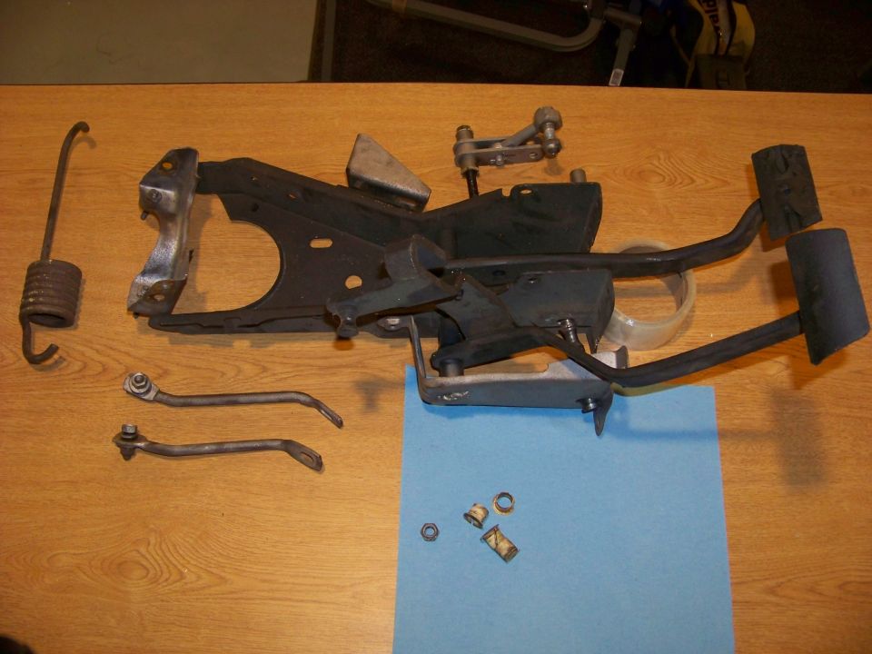 Vehicle pedal assembly with brake and clutch pedals, spring, and linkage parts laid out on a wooden surface