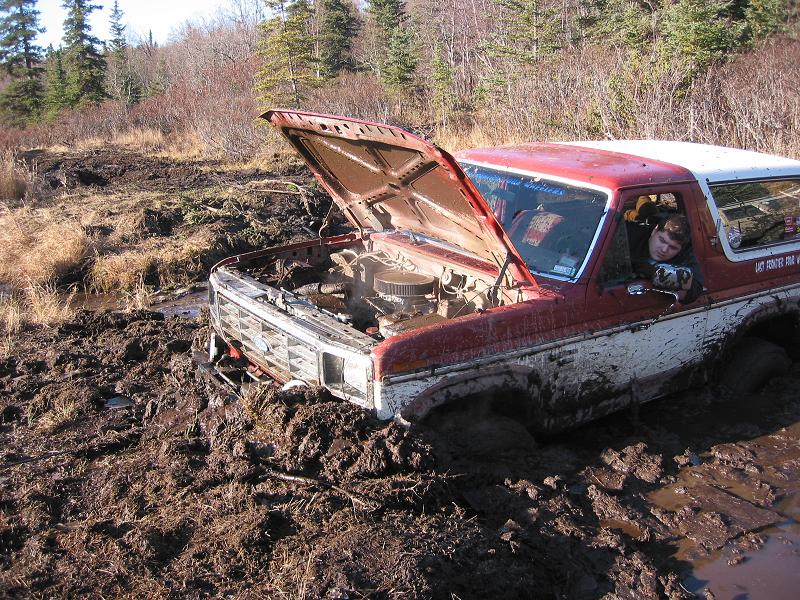 Red and white Ford Bronco stuck deep in mud with hood open, driver inside.