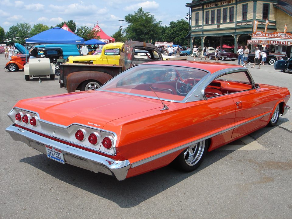 Orange classic car with quad taillights and chrome trim, parked at an outdoor event with other vehicles and buildings in the background