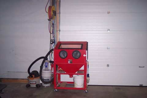 1990 Ford Bronco - A red sandblaster cabinet is set up in a garage next to a shop vacuum and a roll-up door.