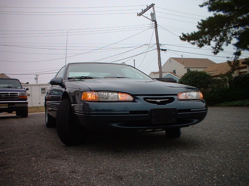 Low front view of a dark green Ford Thunderbird coupe with amber turn signals on, parked on asphalt