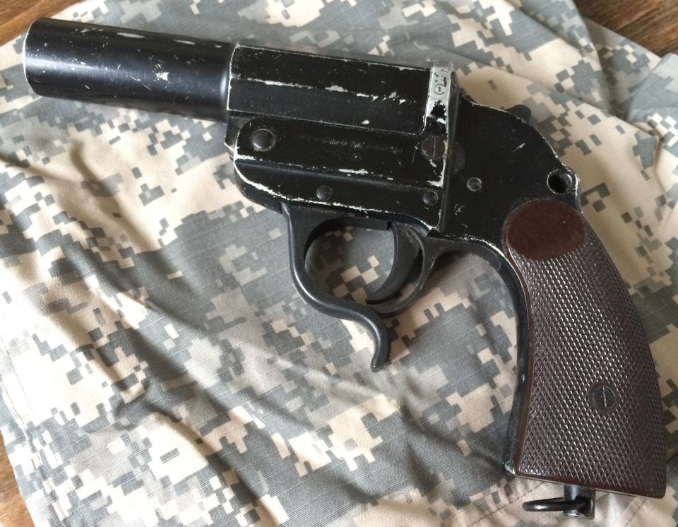 Black flare gun with brown textured grip, showing wear and scratches, resting on camouflage fabric