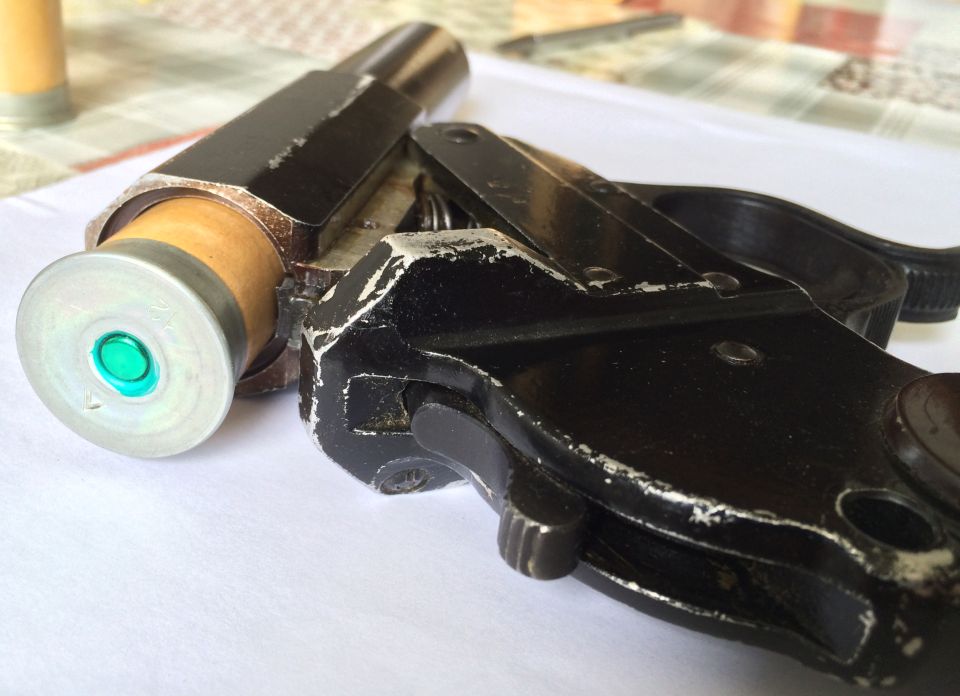 Close-up of a black flare gun loaded with a cartridge. The cartridge has a silver rim and a green primer