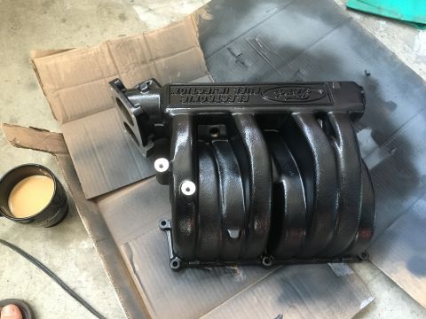1987 Ford Bronco - A freshly painted black Ford intake manifold sits on cardboard next to a cup of coffee.