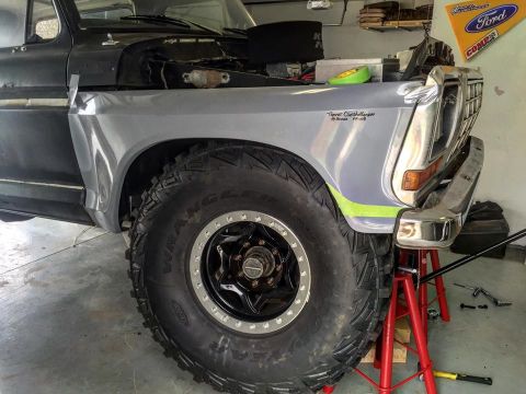 1979 Ford Bronco - The front fender and massive off-road tire of a 1979 Ford Bronco are visible, supported by red jack stands in a garage setting.