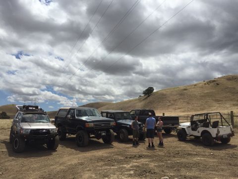 1989 Ford Bronco - A group of modified off-road vehicles are gathered on a dry, grassy hillside under a cloudy sky.