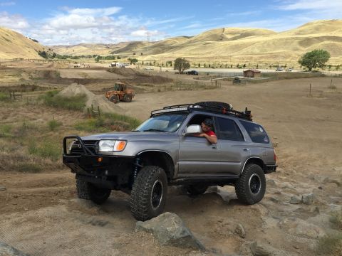 1989 Ford Bronco - A modified silver SUV navigates a rocky trail under a partly cloudy sky.
