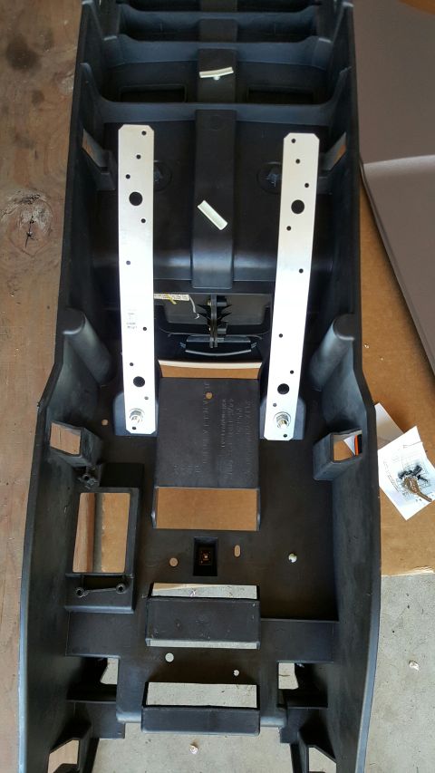 1996 Ford Bronco - Black plastic center console shell removed from the vehicle and lying upside down on a garage floor, with two long drilled aluminum mountin…