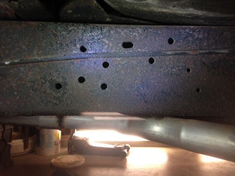 1979 Ford Bronco - Close-up view of a rusted vehicle frame, showing multiple holes and an exhaust pipe.